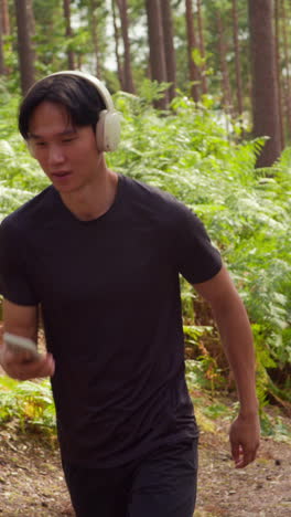 Vertical-Video-Of-Man-Wearing-Wireless-Headphones-Exercising-Doing-Work-Out-Outdoors-Streaming-Music-Or-Podcast-From-Mobile-Phone-Running-Along-Track-In-Forest-Wearing-Sports-Clothing-Shot-In-Real-Time